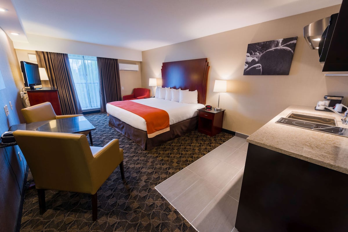 A hotel room with a bed, a desk, a chair, a TV, a painting, and a kitchen area.