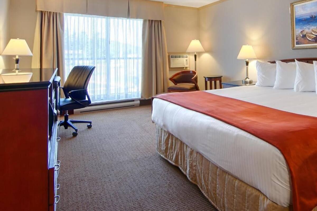 A hotel room with a bed, desk, chair, lamps, and a painting on the wall.