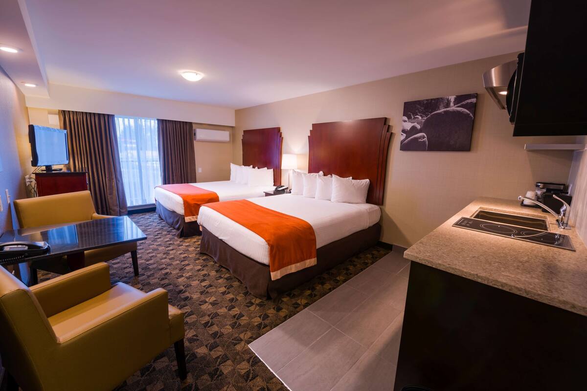 A hotel room with two beds, a desk, a chair, a TV, and a kitchenette.