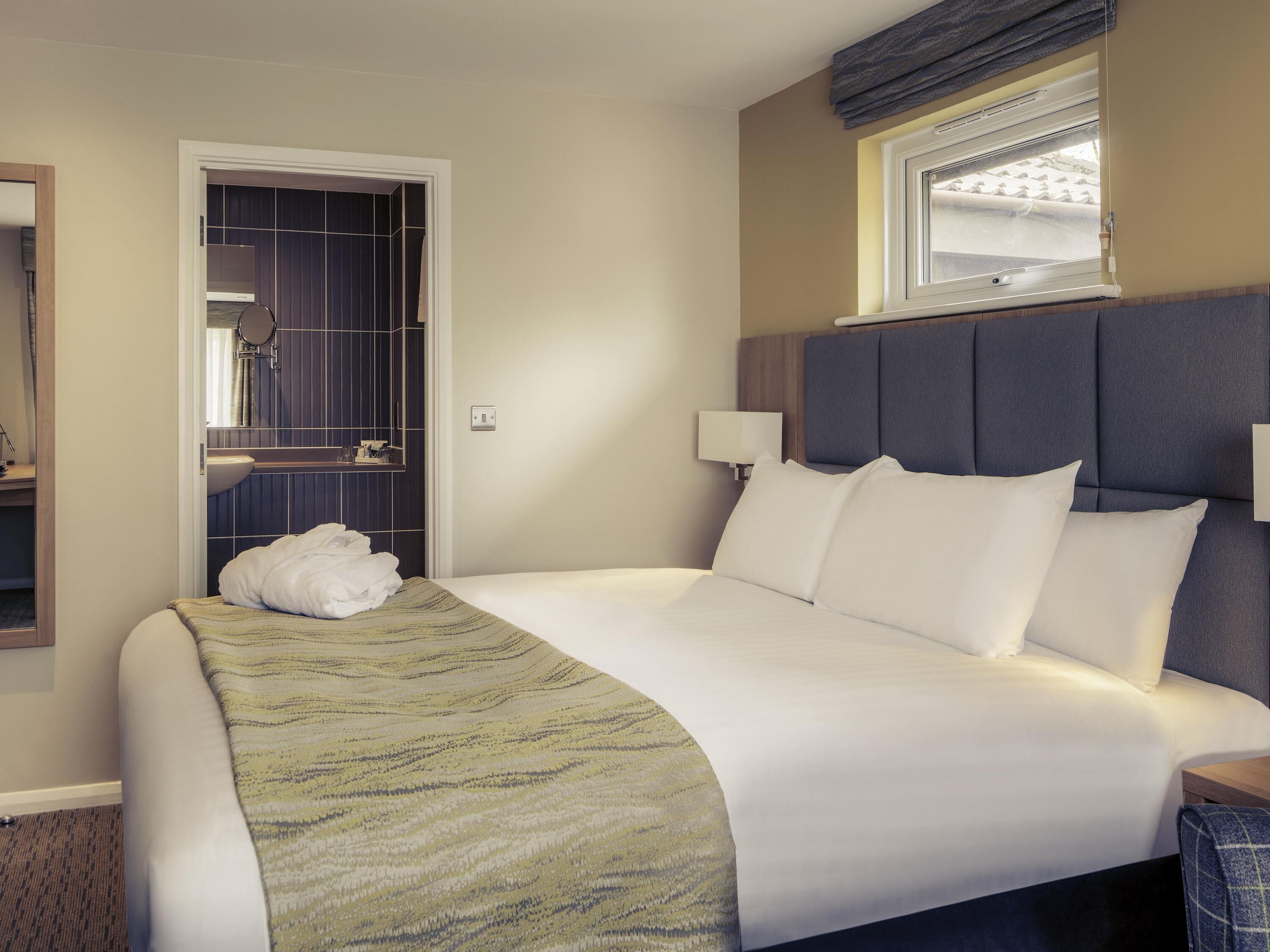 A hotel room with a bed, bedside lamps, a window, a bathroom with a mirror, and a towel on the bed.