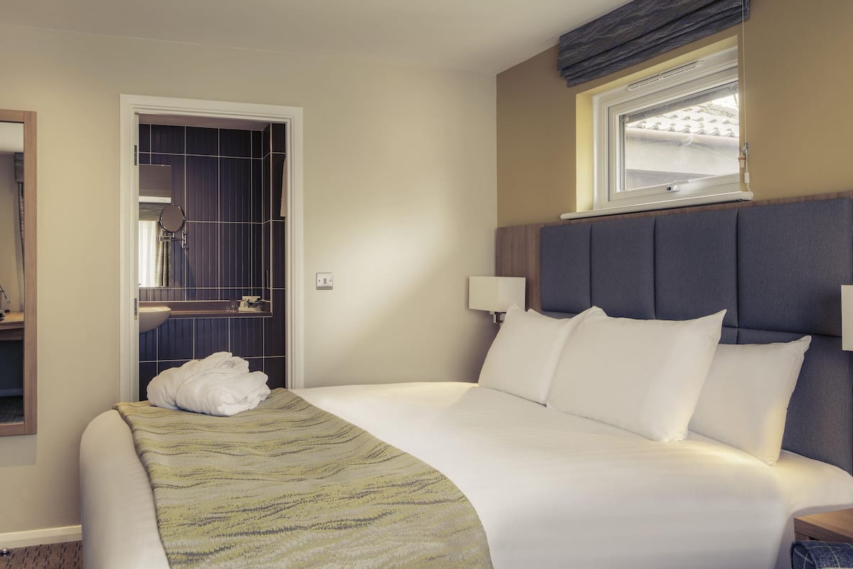 A hotel room with a bed, bedside lamps, a window, a bathroom with a mirror, and a towel on the bed.