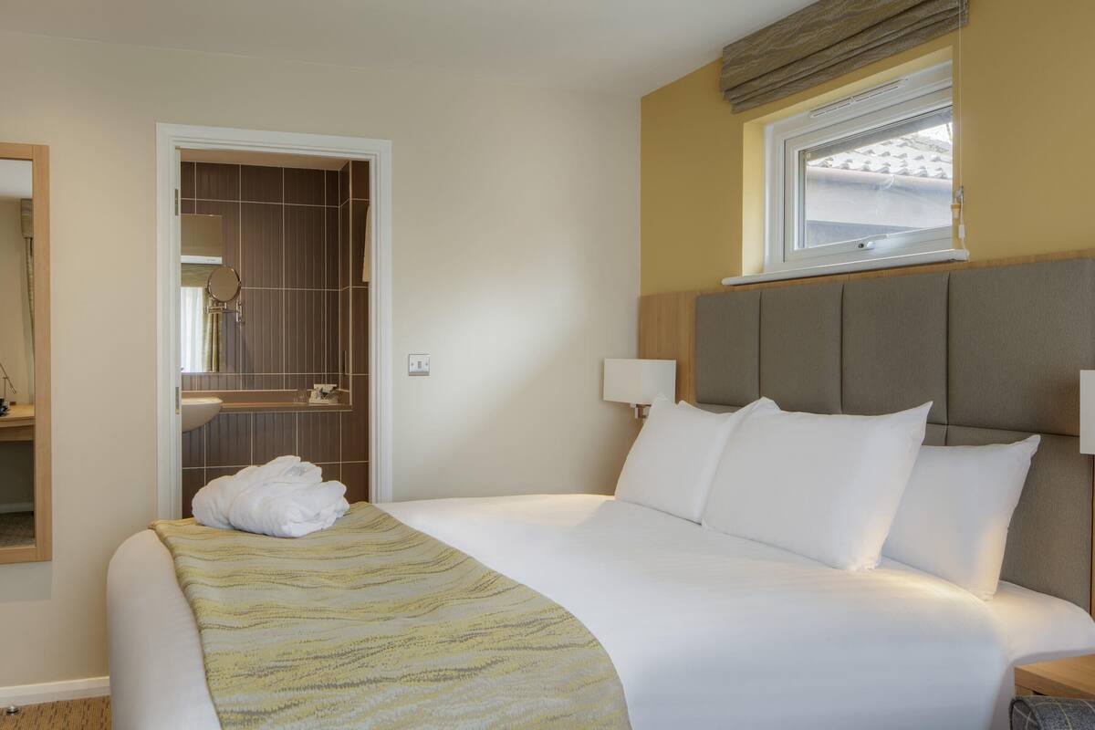 A hotel room with a bed, a bedside table, a lamp, a window with blinds, a mirror, and a bathroom visible through an open door.