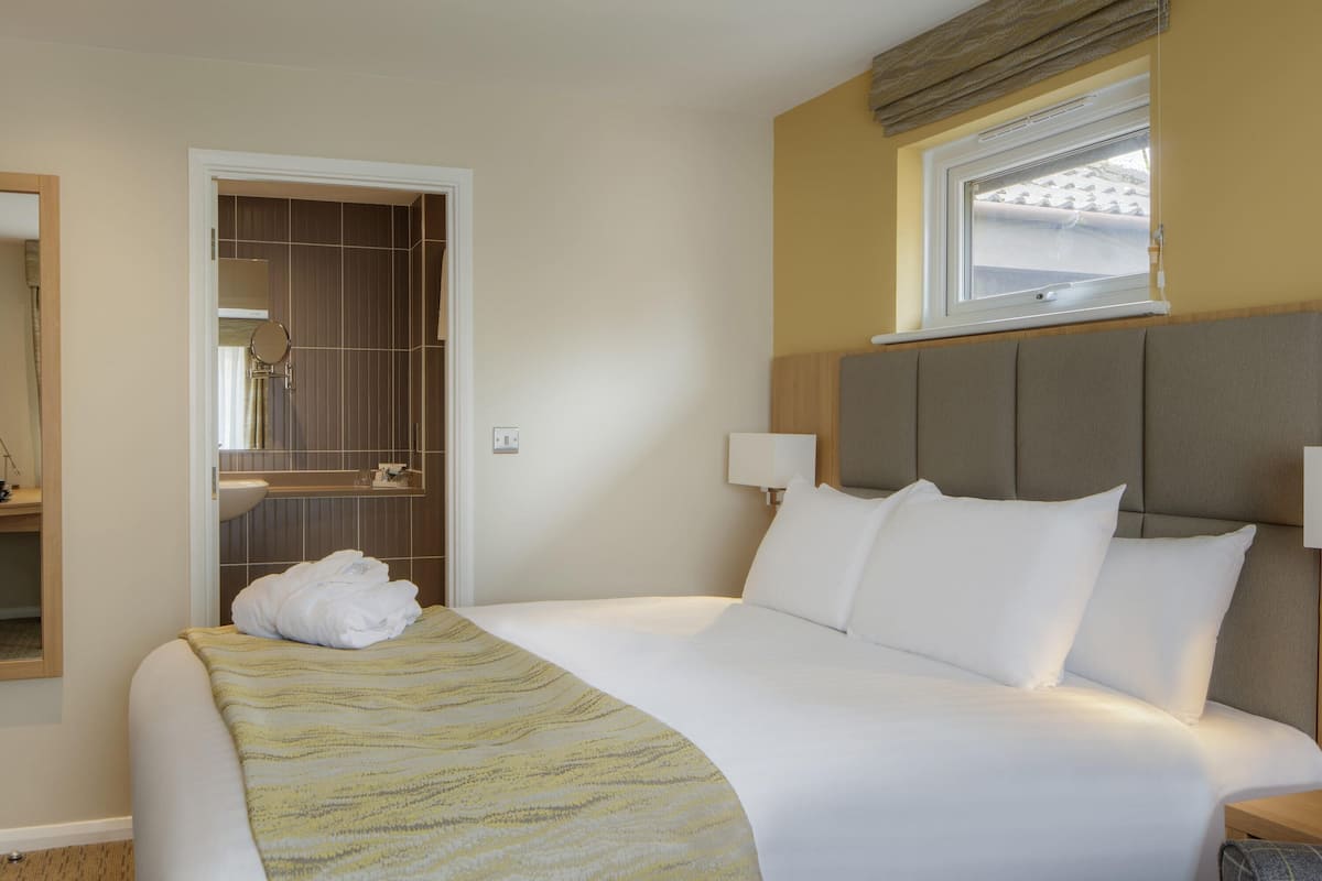 A hotel room with a bed, a bedside table, a lamp, a window with blinds, a mirror, and a bathroom visible through an open door.