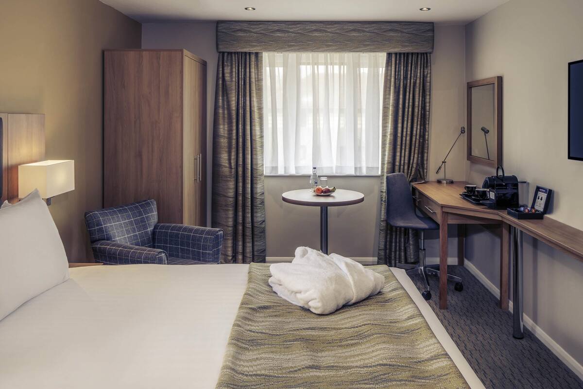 A hotel room with a bed, a desk, a chair, a small table, a lamp, and a window with curtains.