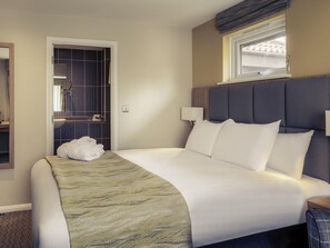 Habitación de hotel con cama, lámparas de noche, ventana y baño con espejo. Hay una toalla sobre la cama.