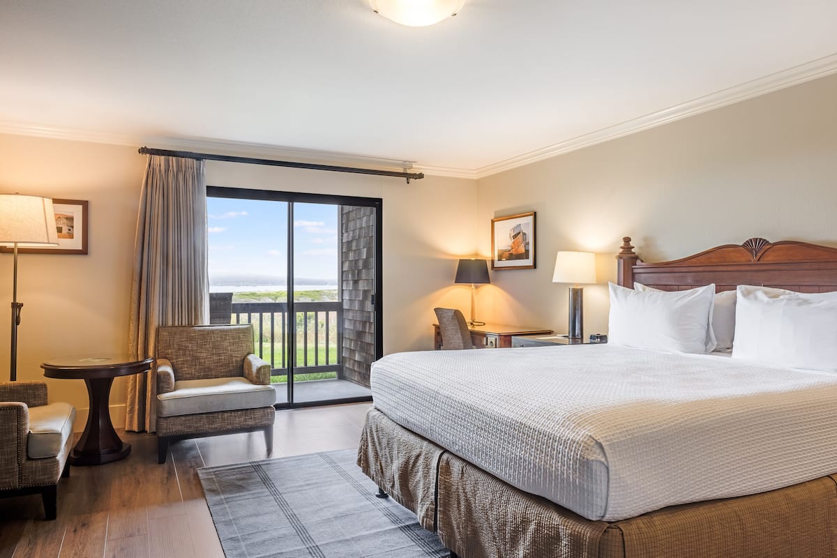 A hotel room with a large bed, a chair, a small table, and a view of the outdoors.