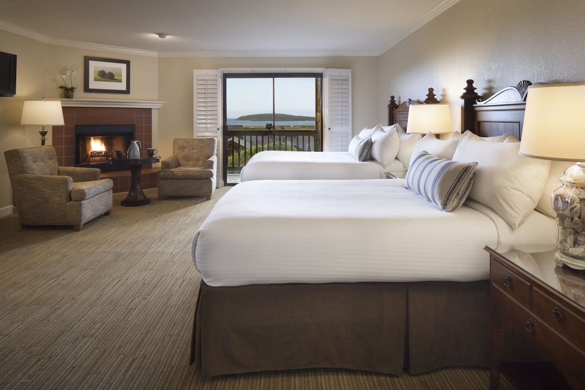 A hotel room with two beds, a fireplace, a balcony with a view, and a seating area with chairs.