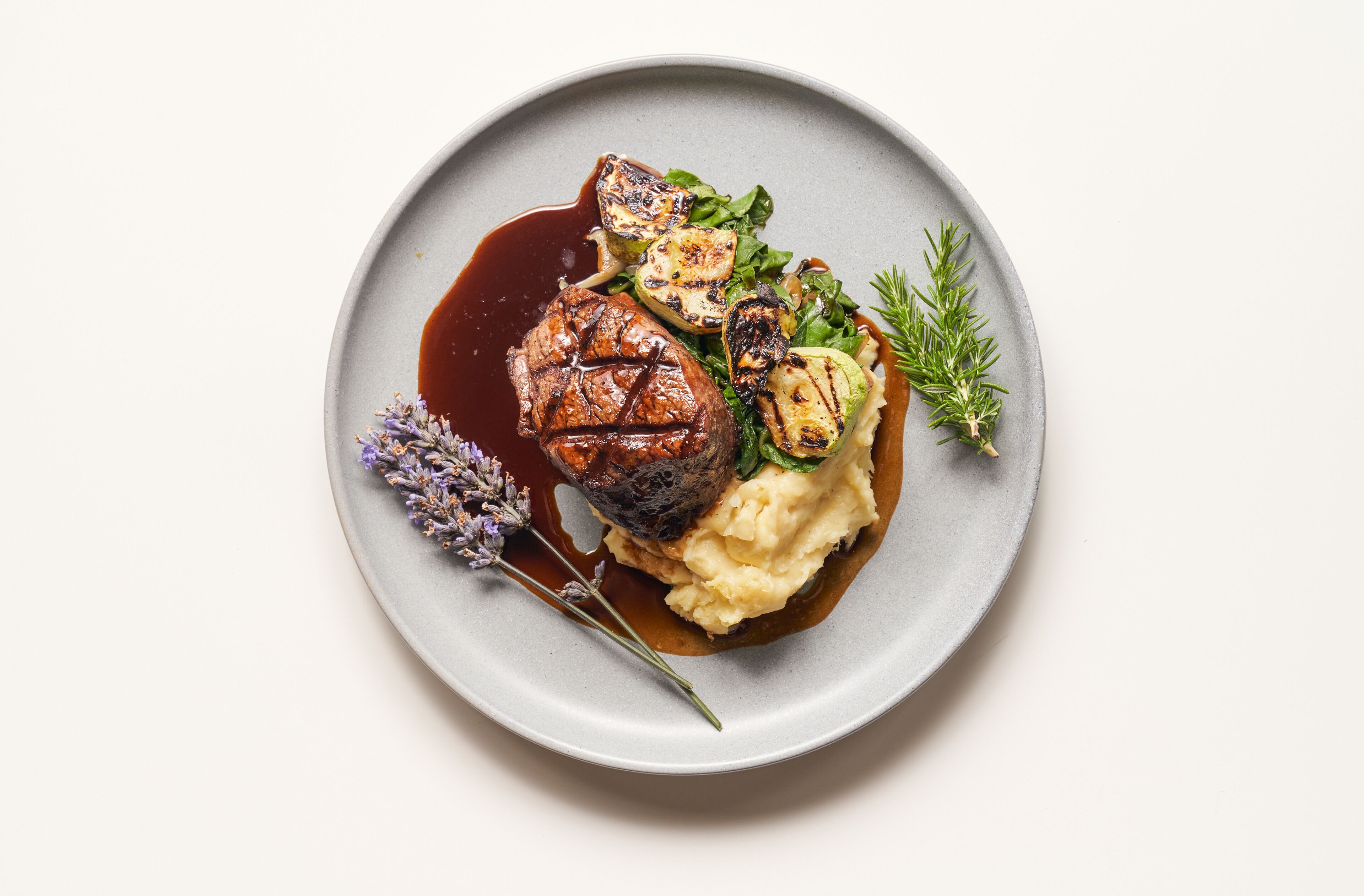 A plate with a steak, grilled vegetables, mashed potatoes, and a sprig of rosemary.