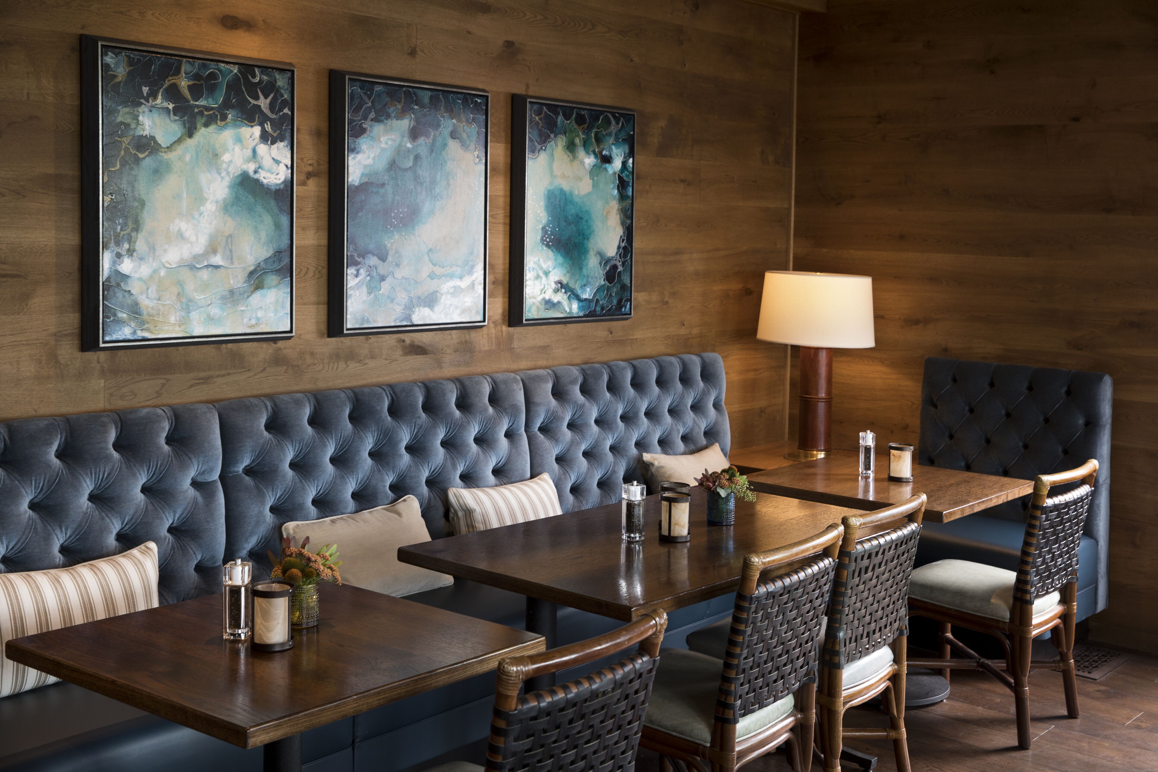 A dining area with a tufted blue sofa, wooden tables, wicker chairs, and abstract artwork on the wall.