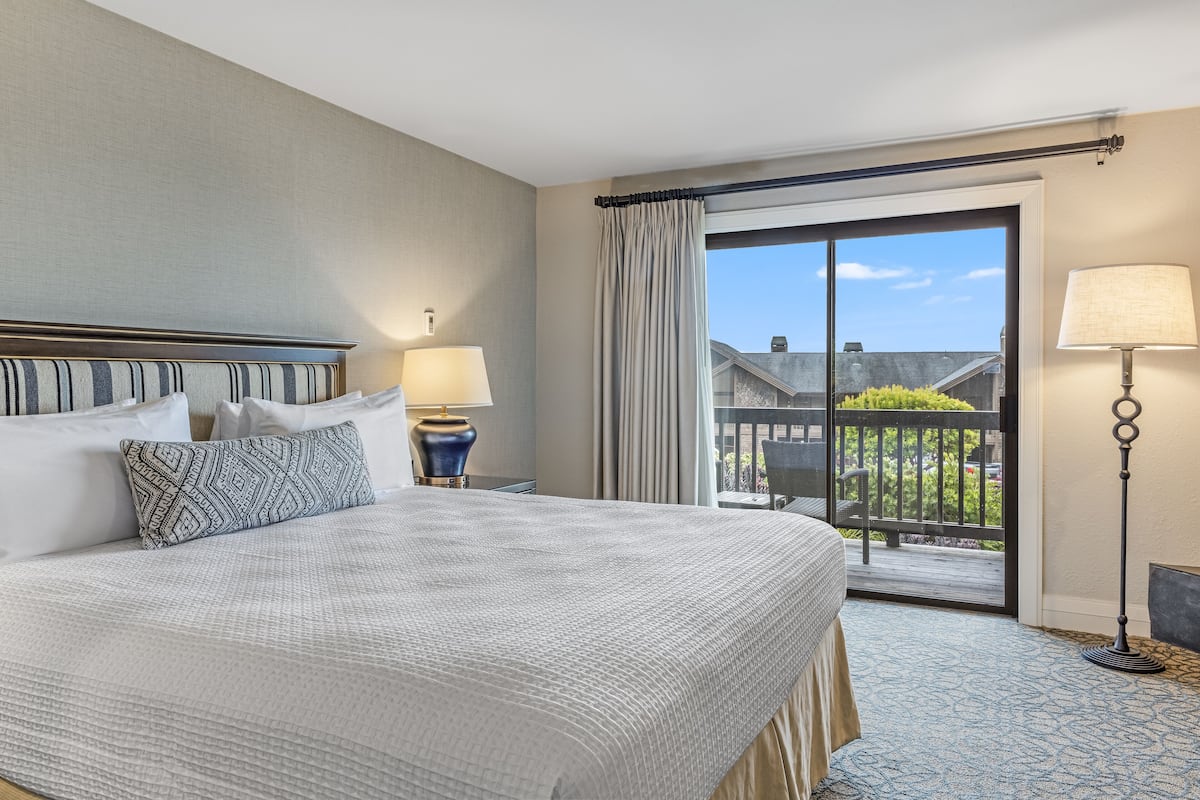 A hotel room with a large bed, a nightstand with a lamp, a view of the outside balcony, and a window with curtains.