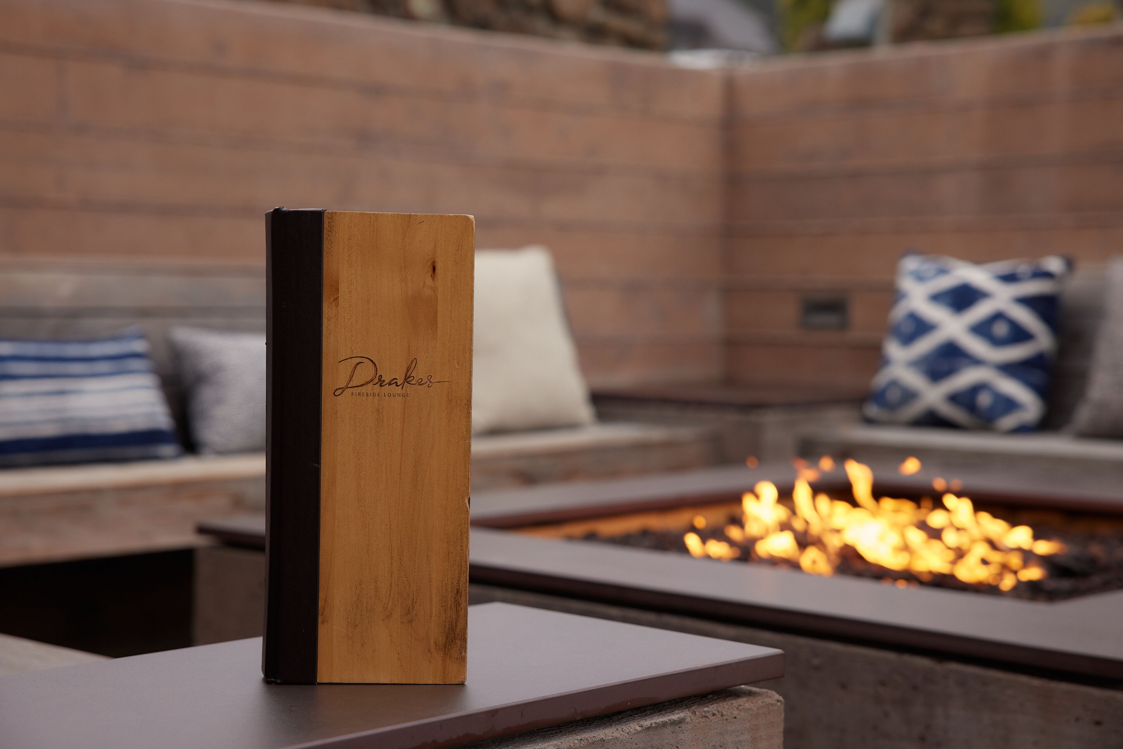 Una caja de madera con la inscripción “Drakes” sobre una mesa en un área de estar al aire libre acogedora, con una chimenea.
