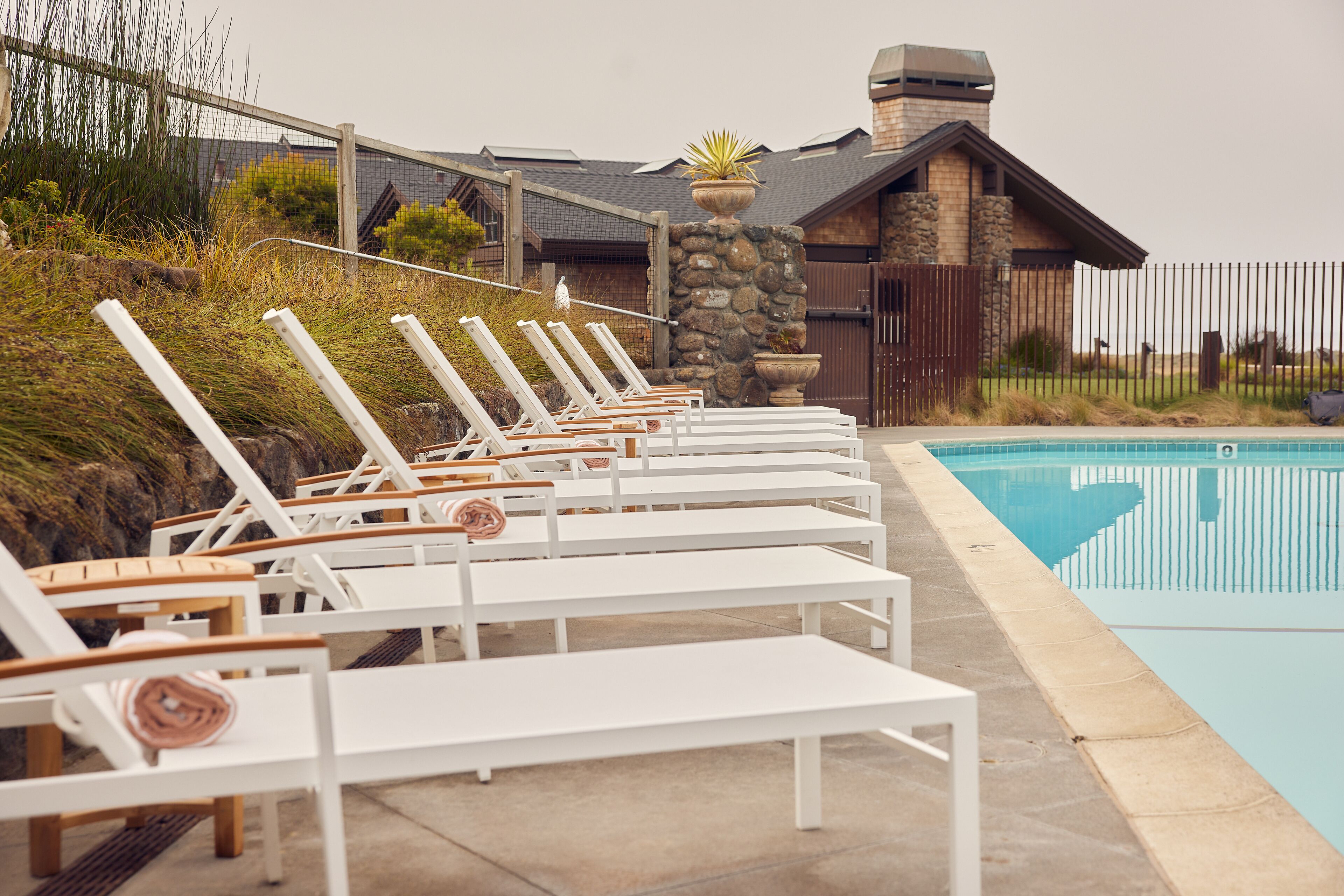 A row of white lounge chairs with wooden seats and backs, arranged along a poolside.