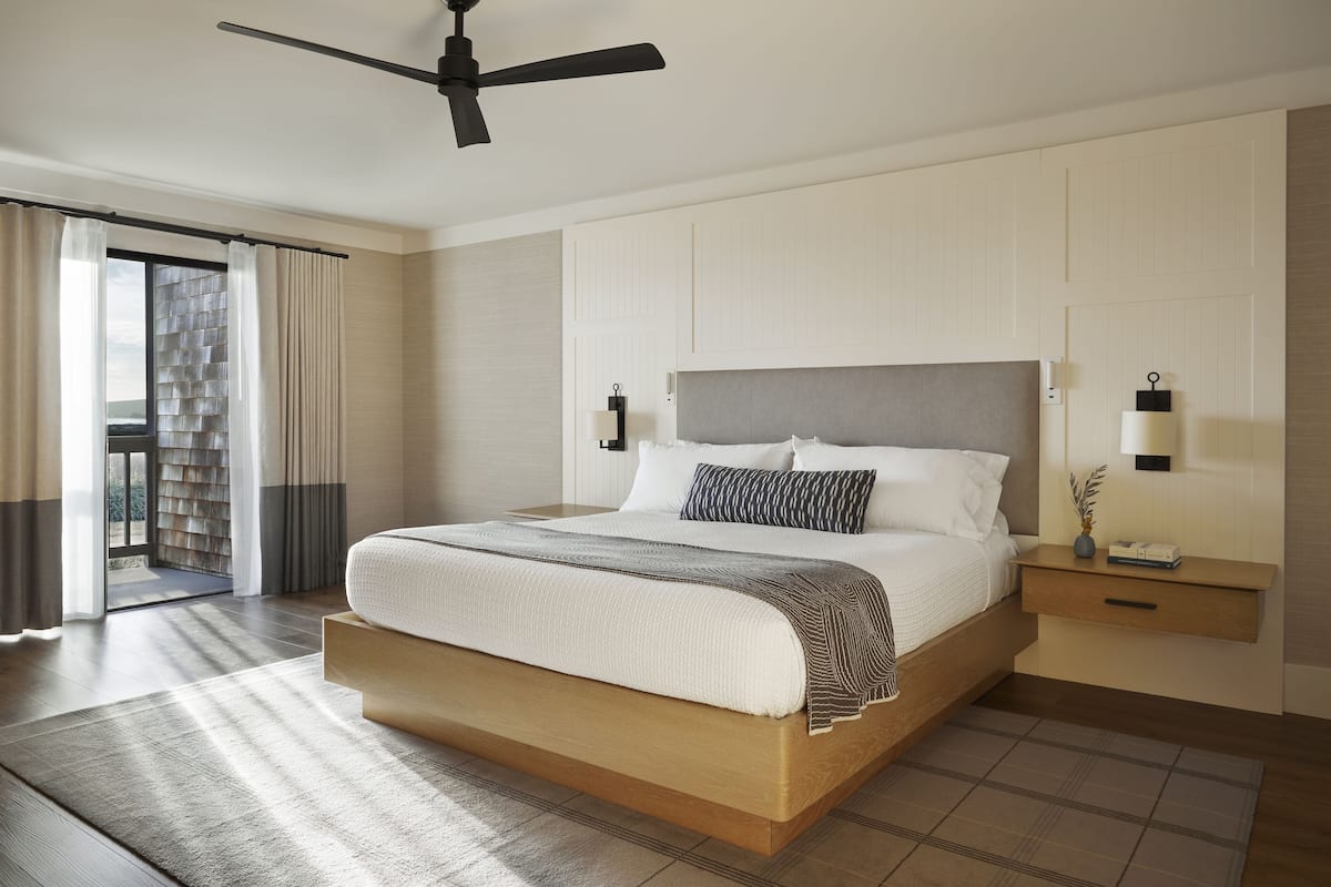 A modern hotel bedroom with a large bed, a ceiling fan, a nightstand with a lamp, and a view of the outdoors.