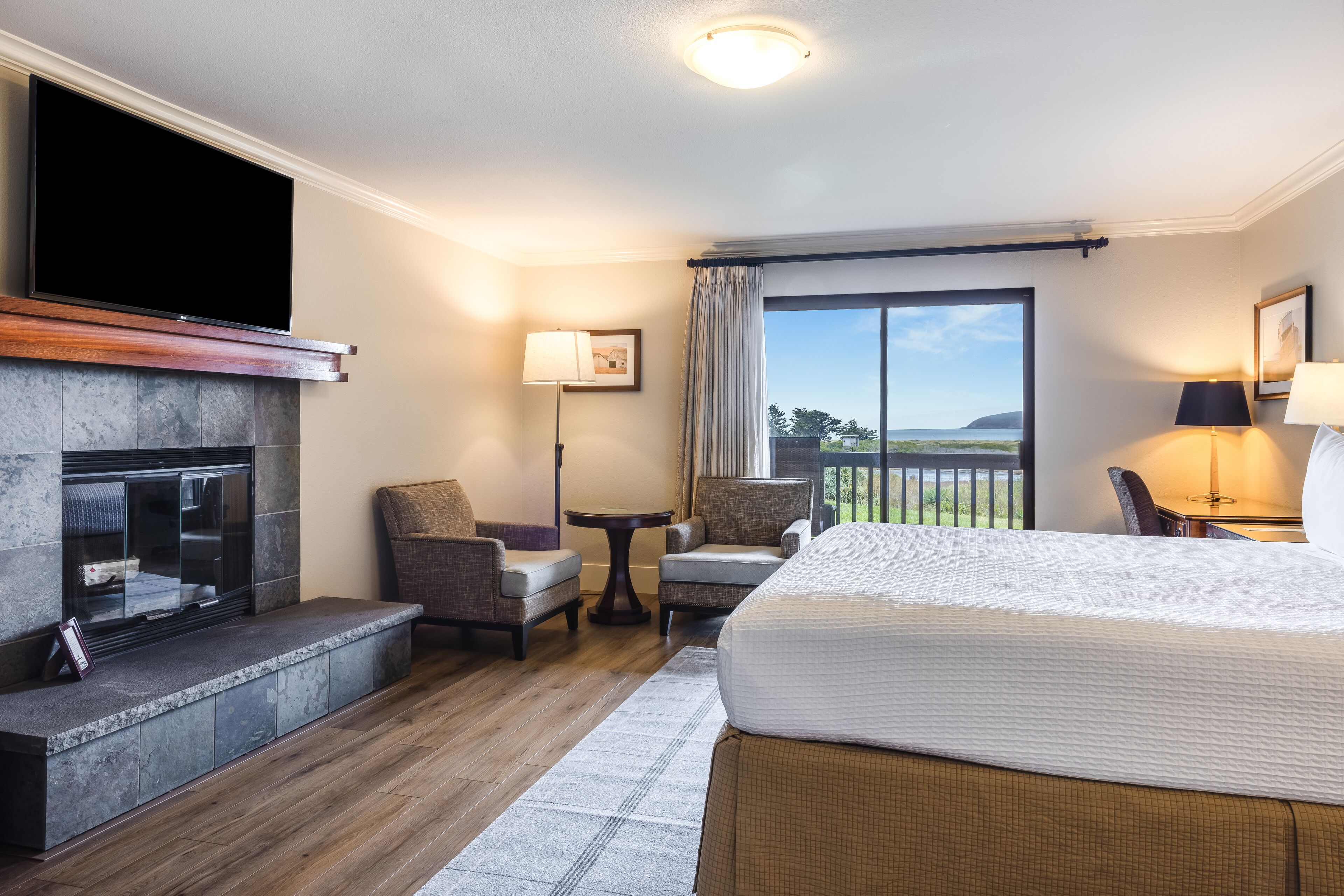 A hotel room with a large bed, a fireplace, two armchairs, a small table, and a view of the outdoors.