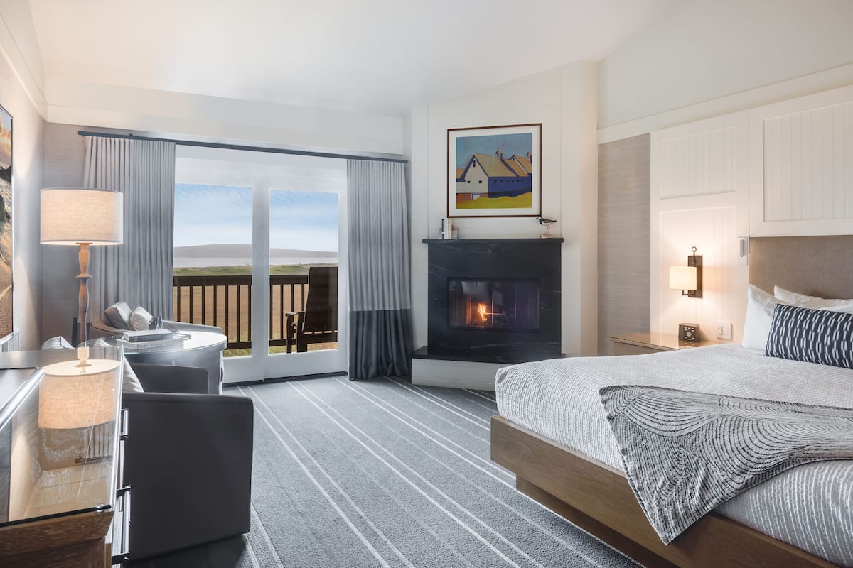 A hotel room with a bed, a desk, a chair, a fireplace, and a large window with curtains.