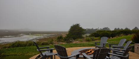 Un pozo de fuego circular con cuatro sillas Adirondack a su alrededor, con vistas a una playa y al océano.