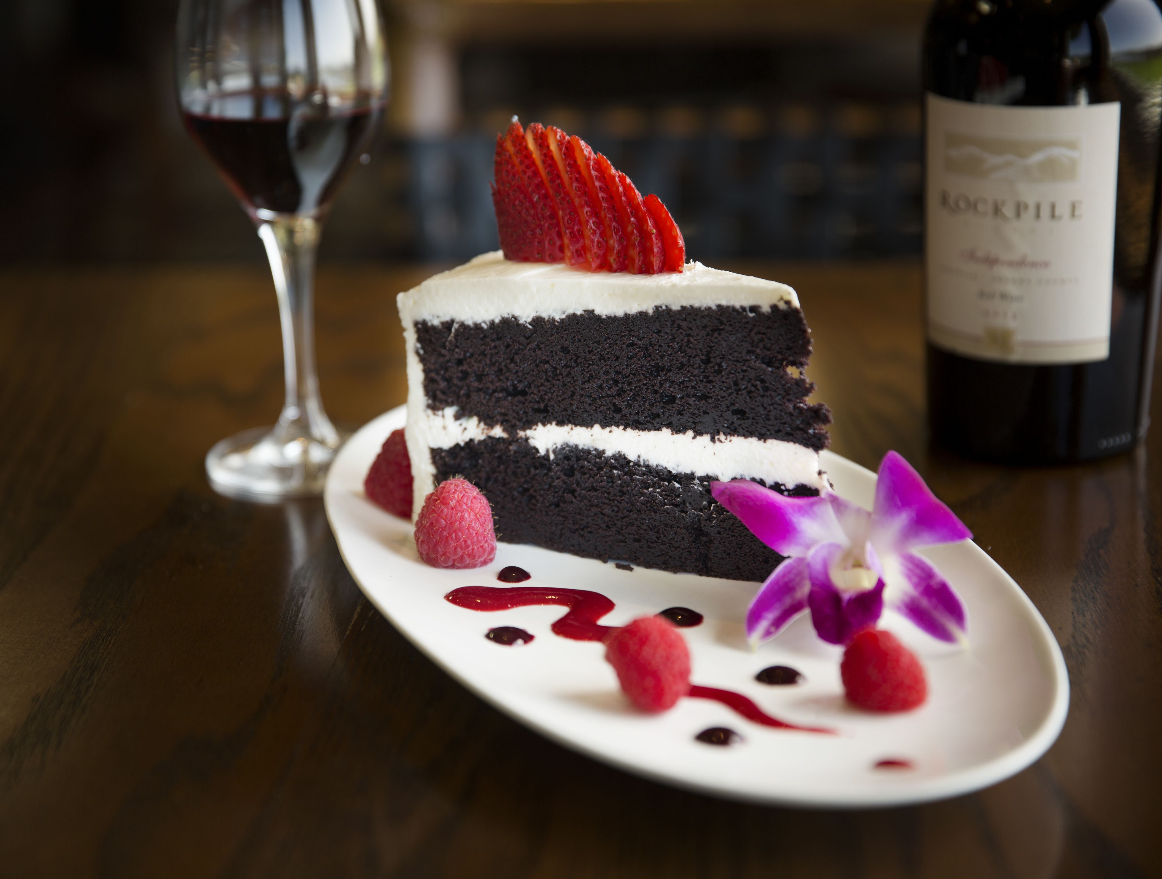 Ein Stück Schichttorte mit Erdbeeren und Himbeeren auf einem weißen Teller, dazu eine Weinflasche und ein Weinglas mit Rotwein.