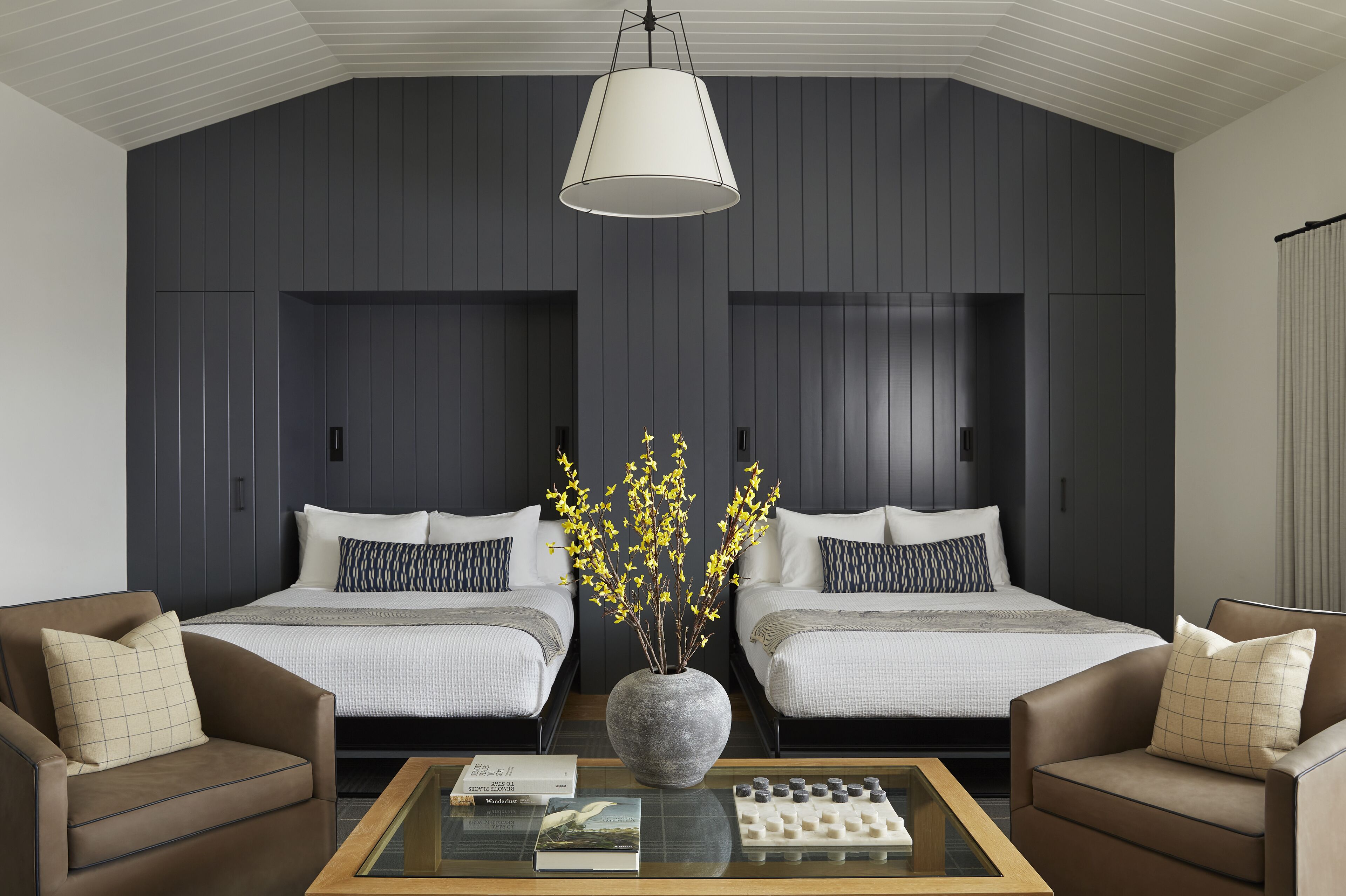 Une chambre d’hôtel moderne avec deux lits, un grand vase rempli de fleurs jaunes et une table basse avec des livres et un jeu d’échecs.
