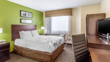 Ein Hotelzimmer mit einem großen Bett, einem Schreibtisch mit Fernseher und einem Sessel. An der Wand hängen zwei gerahmte Bilder, ein Fenster mit Vorhängen und ein Teppichboden.