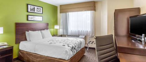 A hotel room with a large bed, a desk with a television, and a chair. There are two framed pictures on the wall, a window with curtains, and a carpeted floor.