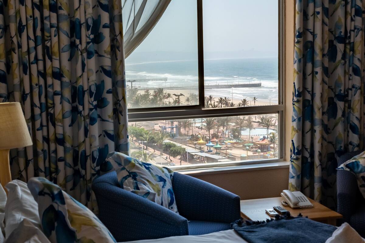 A hotel room with a large window, floral curtains, a bed with blue bedding, and a view of the beach and cityscape.