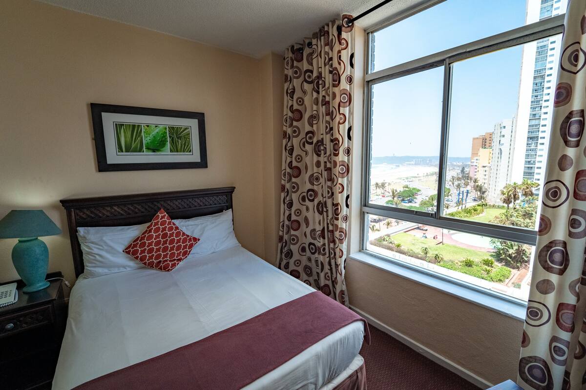 A hotel room with a bed, a nightstand with a lamp, a framed picture on the wall, and a window with a view of buildings and greenery.