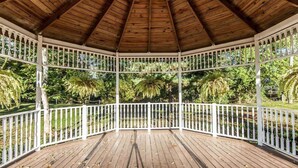 Um gazebo com telhado de madeira e grades brancas, rodeado por vegetação exuberante.