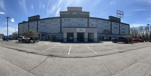A multi-story hotel with a central entrance, parking lot, and a clear sky.
