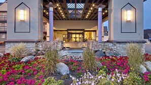 A hotel entrance with a modern design, featuring a stone facade, a welcoming entrance with a glass door, and a landscaped garden with colorful flowers. - Coast Kamloops Hotel & Conference Centre (Kamloops)