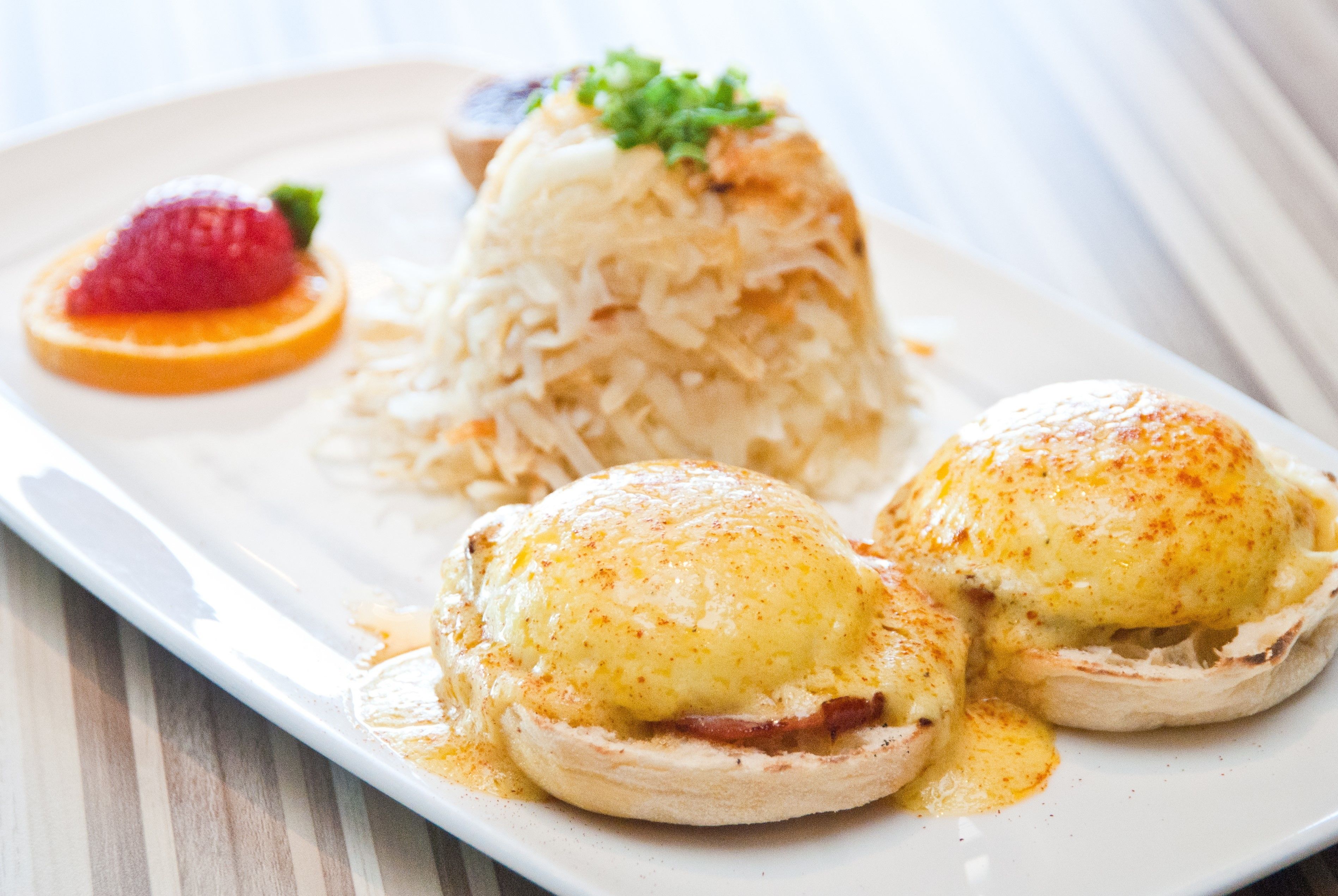 A plate with eggs benedict, a slice of orange, and a raspberry.