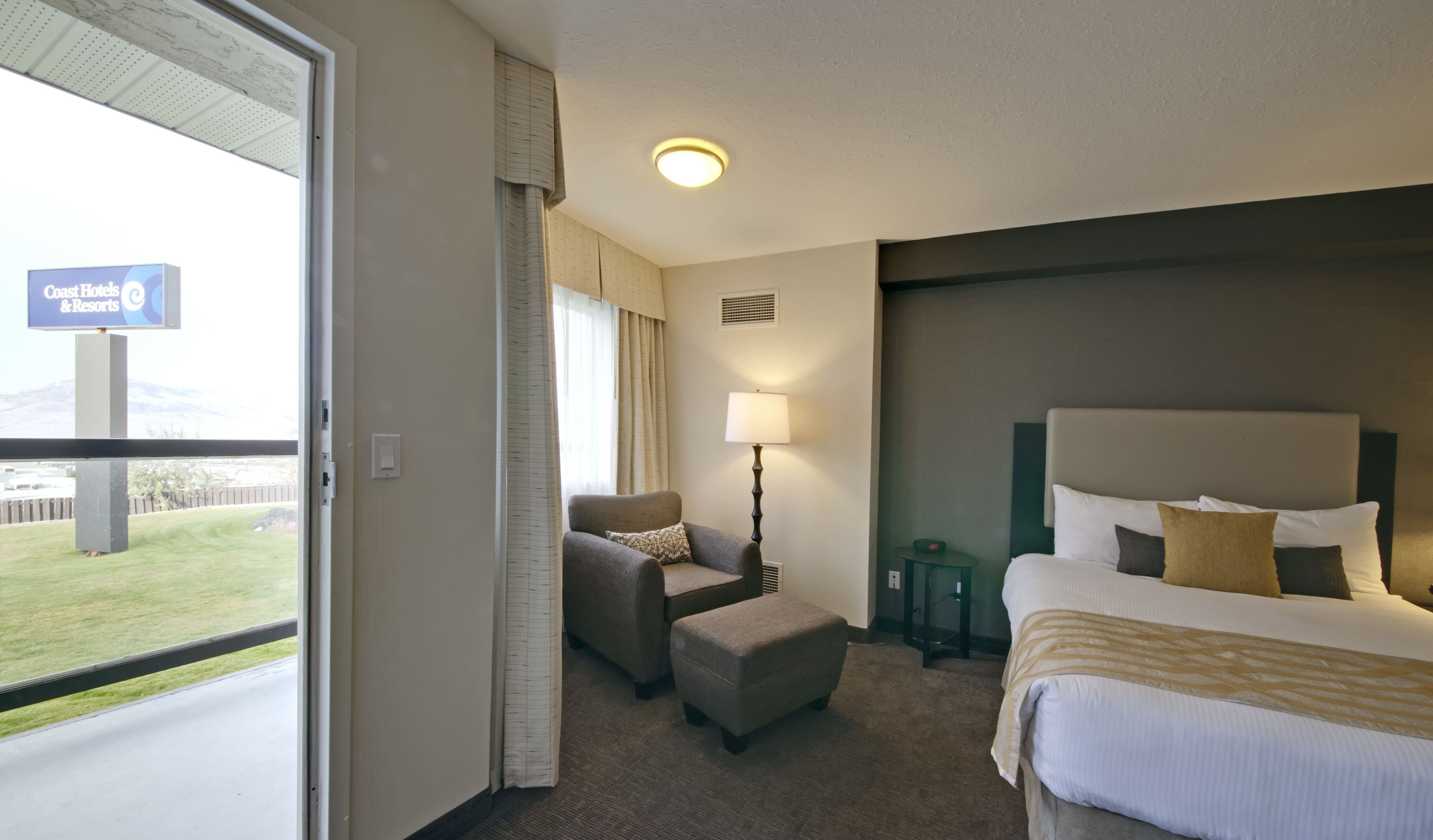 Ein Hotelzimmer mit einem Bett, einem Sessel, einem Hocker, einer Lampe und Blick ins Freie.