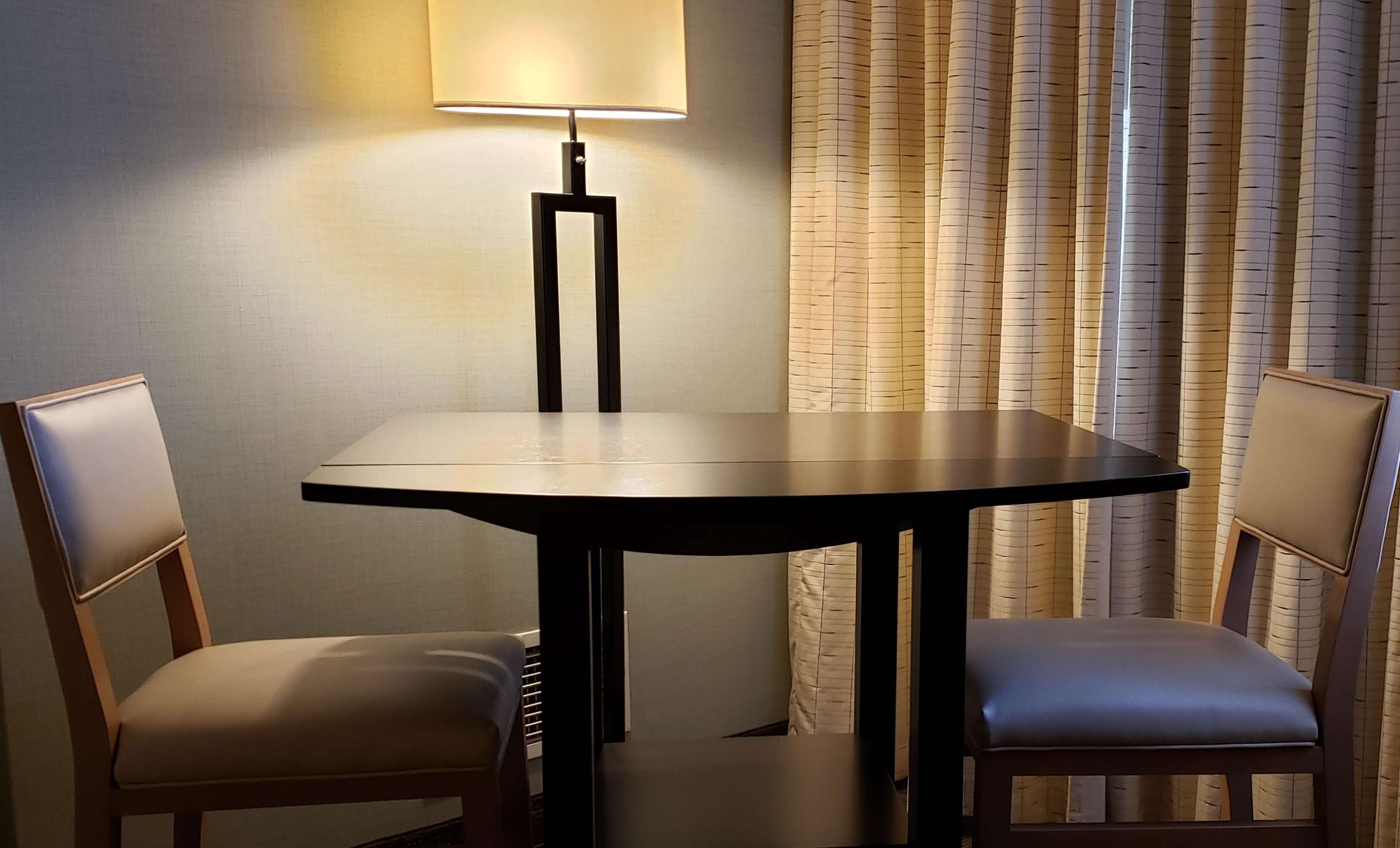 Une petite table ronde avec un cadre noir, deux chaises dotées d’assises rembourrées, un lampadaire et un rideau à motifs verticaux.