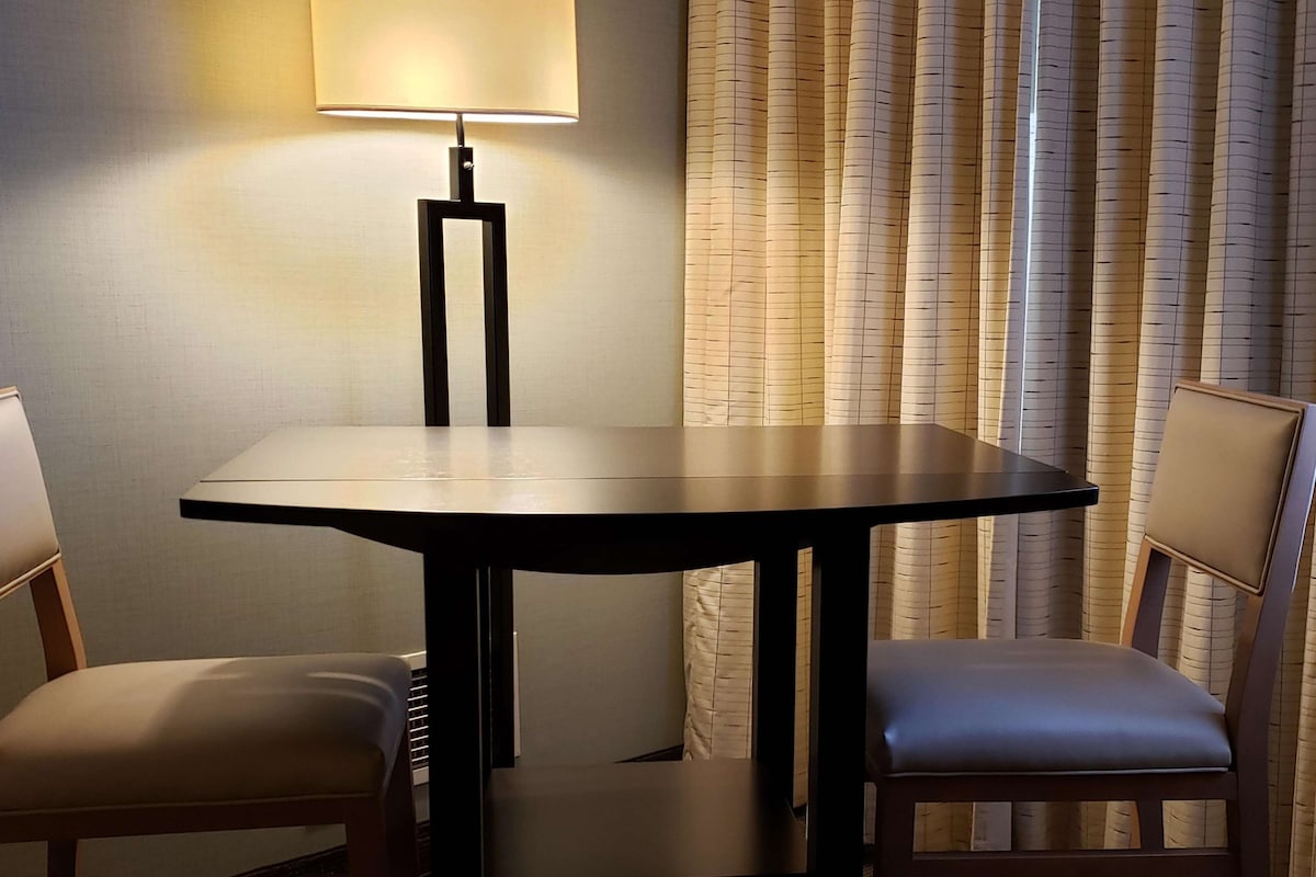 A small, round table with a black frame, two chairs with cushioned seats, a floor lamp, and a curtain with a vertical pattern.