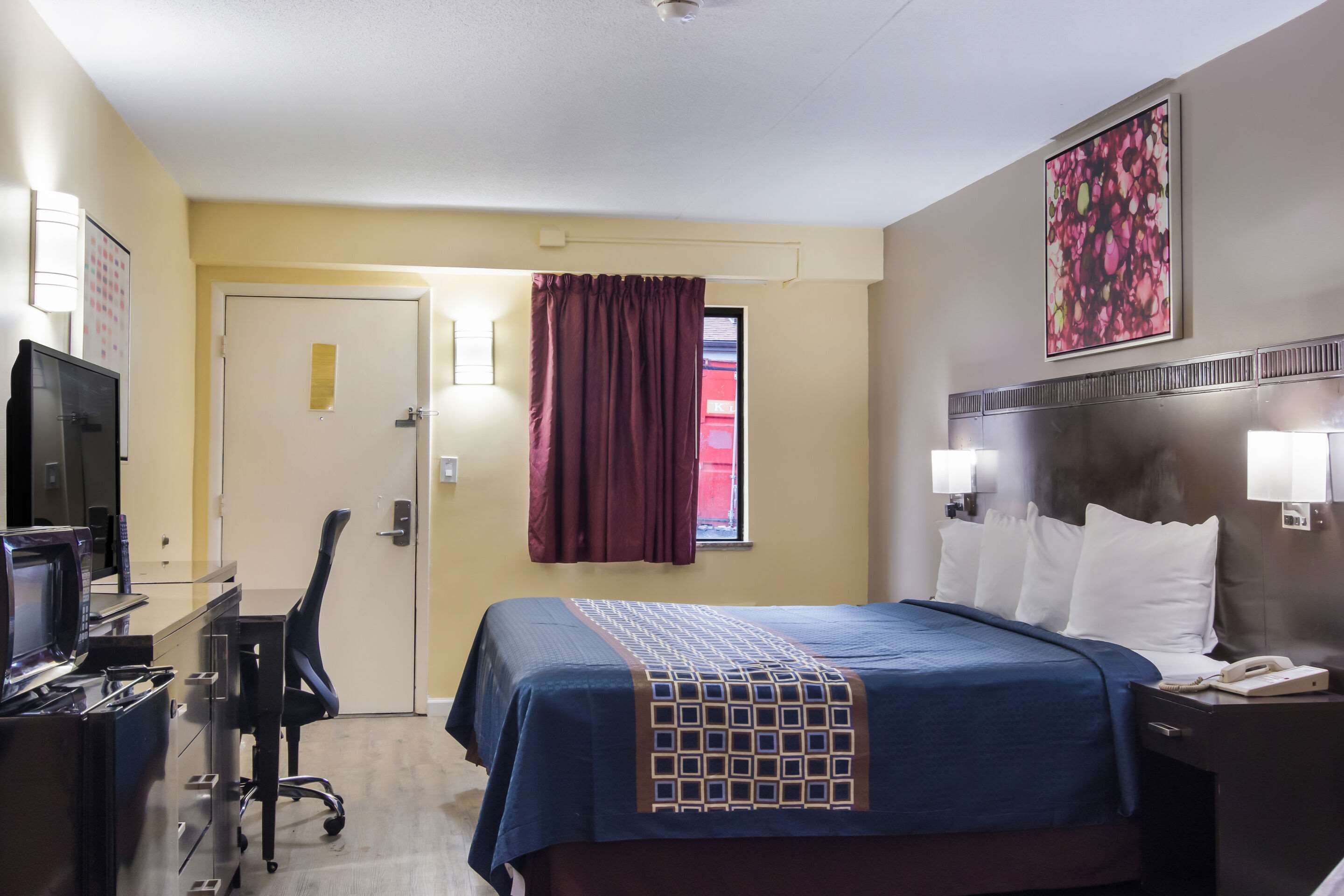 A hotel room with a bed, a desk with a computer, a chair, a TV, and a window with curtains.