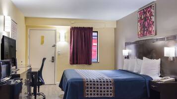 A hotel room with a bed, a desk with a computer, a chair, a TV, and a window with curtains.