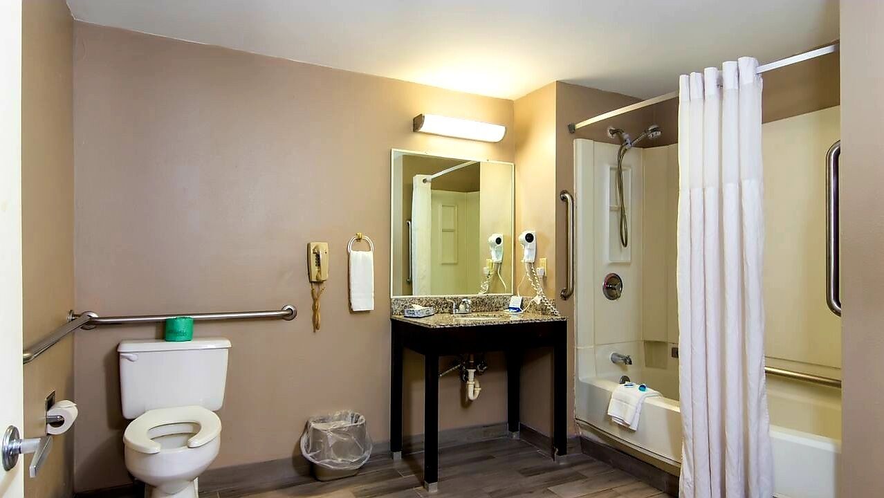 A bathroom with a toilet, sink, mirror, shower, and grab bars.