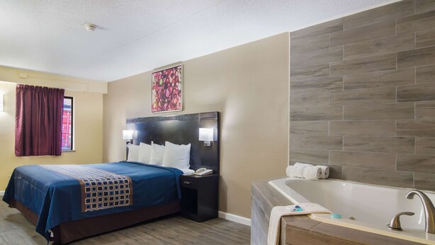 Habitación de hotel con una cama grande, una mesita de noche, un televisor montado en la pared, un baño con bañera y suelo de madera.