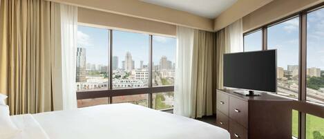 Habitación de hotel con una cama grande, un televisor de pantalla plana, un aparador de madera y vistas a la ciudad a través de amplios ventanales.