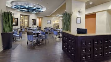A modern hotel lobby with multiple dining tables and chairs, a reception desk, and decorative plants.