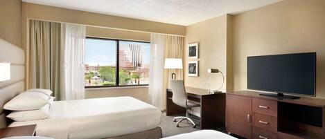 A hotel room with two beds, a desk, a TV, and a window with a view of buildings.
