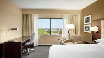 Habitación de hotel con una cama grande, un escritorio, una silla, una mesita pequeña y una mesita de noche con lámpara. Se ve una ventana con cortinas y vistas a edificios.