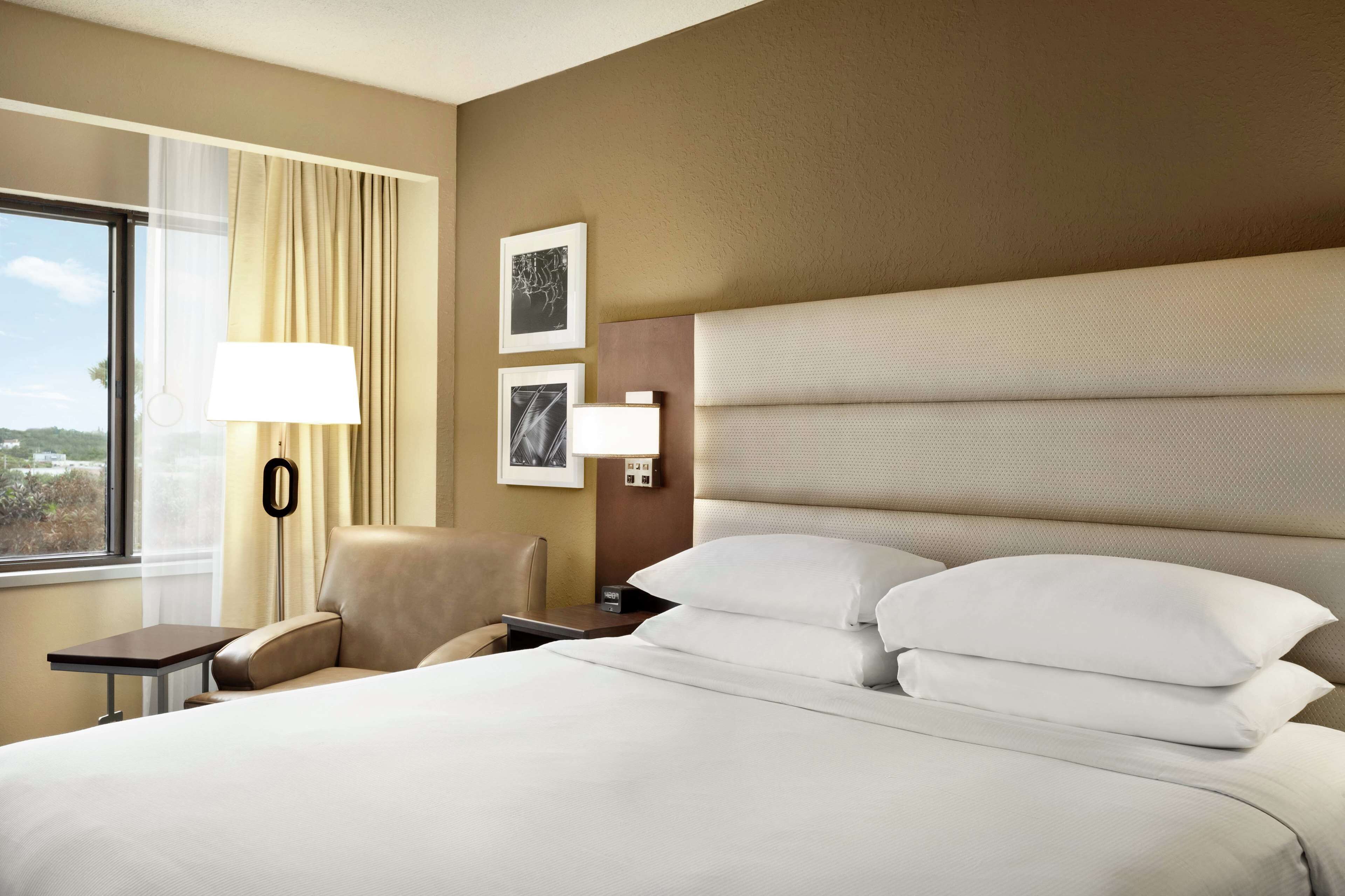 Een hotelkamer met een groot bed, nachtkastje, lamp, stoel en raam met gordijnen.