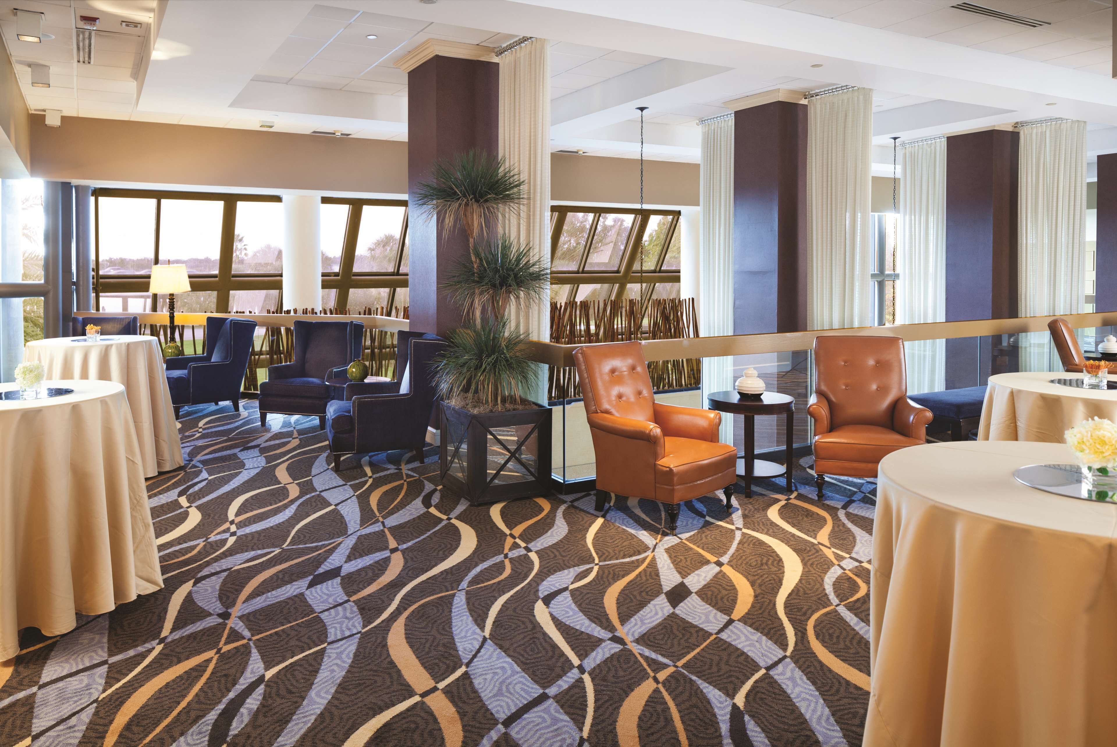 A hotel lobby with round tables, leather chairs, a large potted plant, and large windows offering a view.