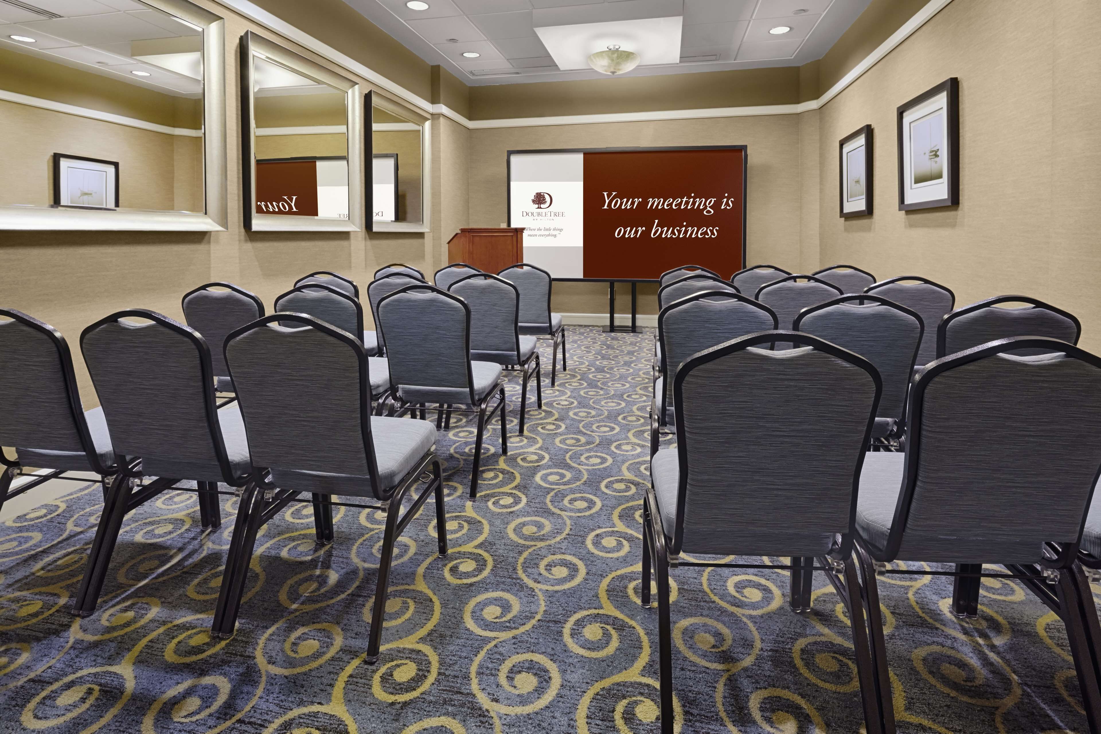 Un salón de conferencias con filas de sillas, un atril y una pantalla de presentación que muestra el texto 'Your meeting is our business'.