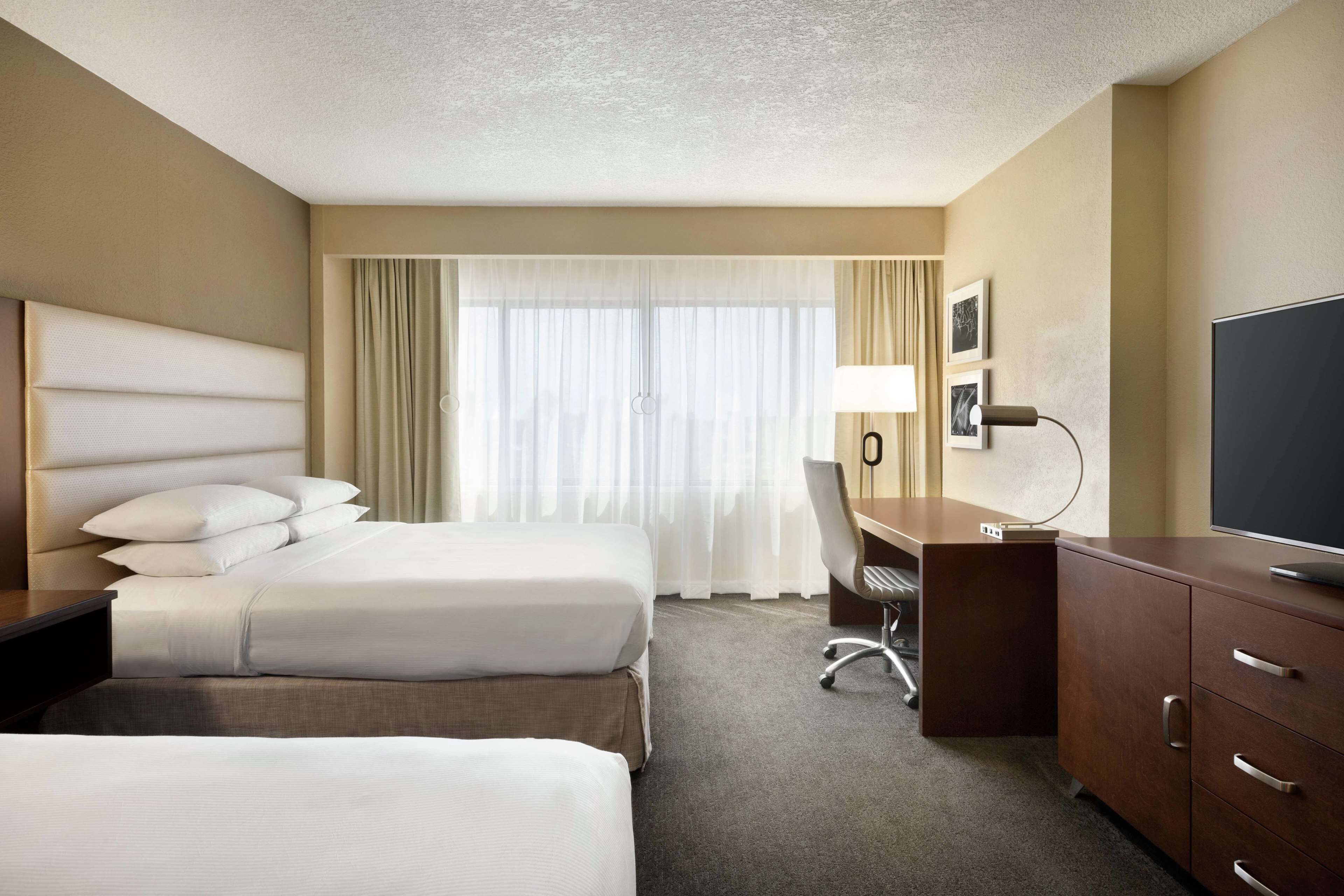 Une chambre d’hôtel avec deux lits, un bureau, une chaise, une télévision et une grande fenêtre avec des rideaux.