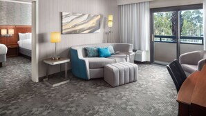 Une chambre d’hôtel avec un canapé gris, un pouf assorti, une petite table, une lampe et une grande fenêtre donnant sur des arbres.