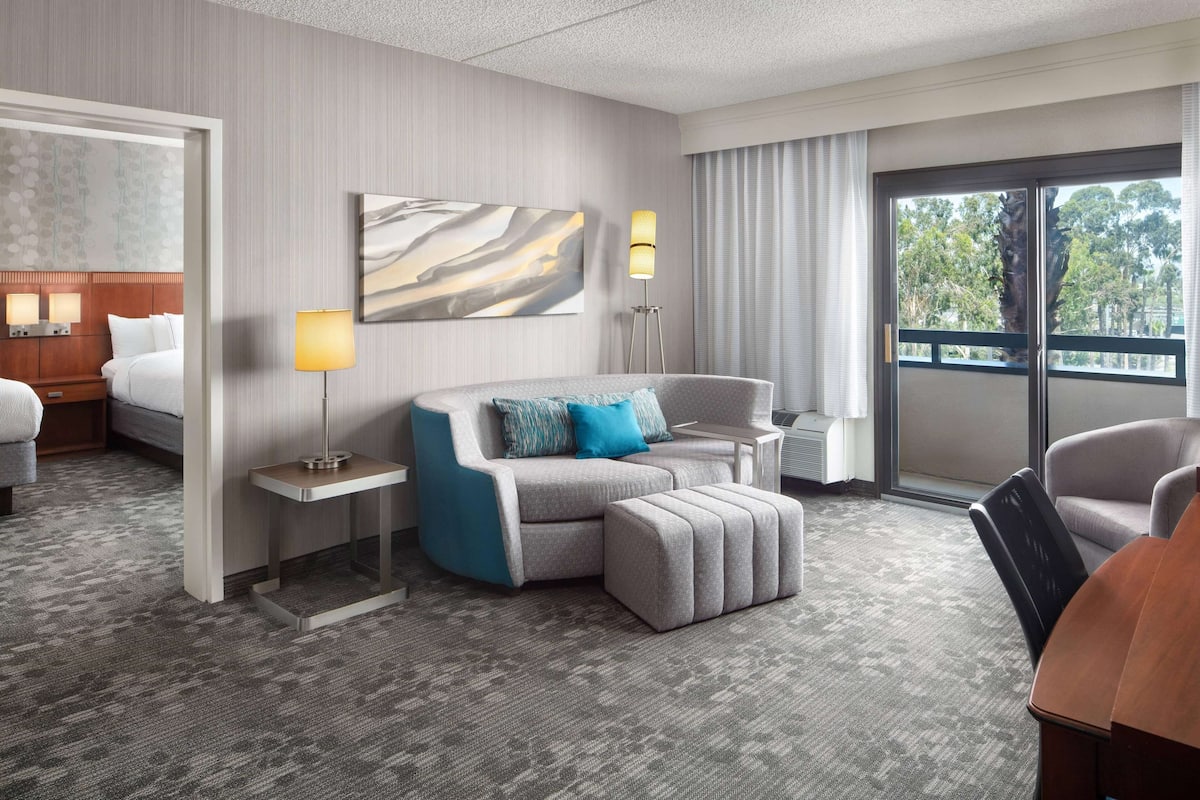 Une chambre d’hôtel avec un canapé gris, un pouf assorti, une petite table, une lampe et une grande fenêtre donnant sur des arbres.