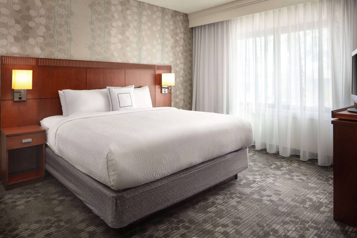 Une chambre d’hôtel avec un grand lit, deux tables de chevet avec des lampes, une télévision et une fenêtre avec des rideaux légers.