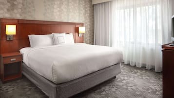 Habitación de hotel con una cama grande, dos mesitas de noche con lámparas, un televisor y una ventana con cortinas translúcidas.