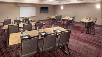 Un salón de conferencias con varias mesas y sillas, equipado con botellas de agua y vasos, una pantalla de proyector y un televisor.